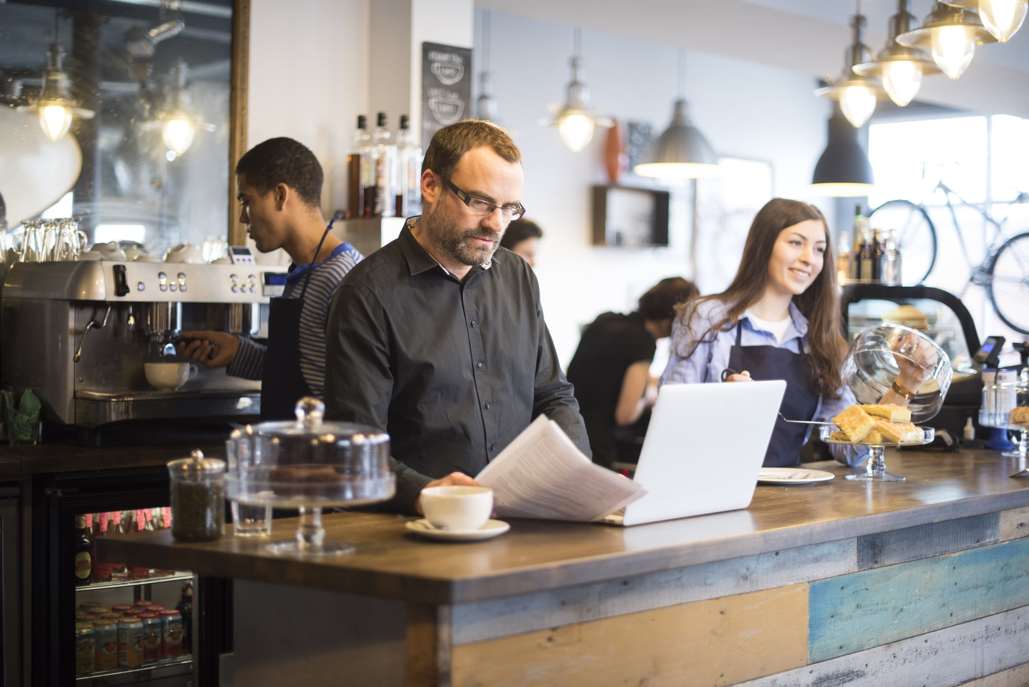 Man leser avisen mens han nyter en kopp kaffe på en café og har laptopen åpen.