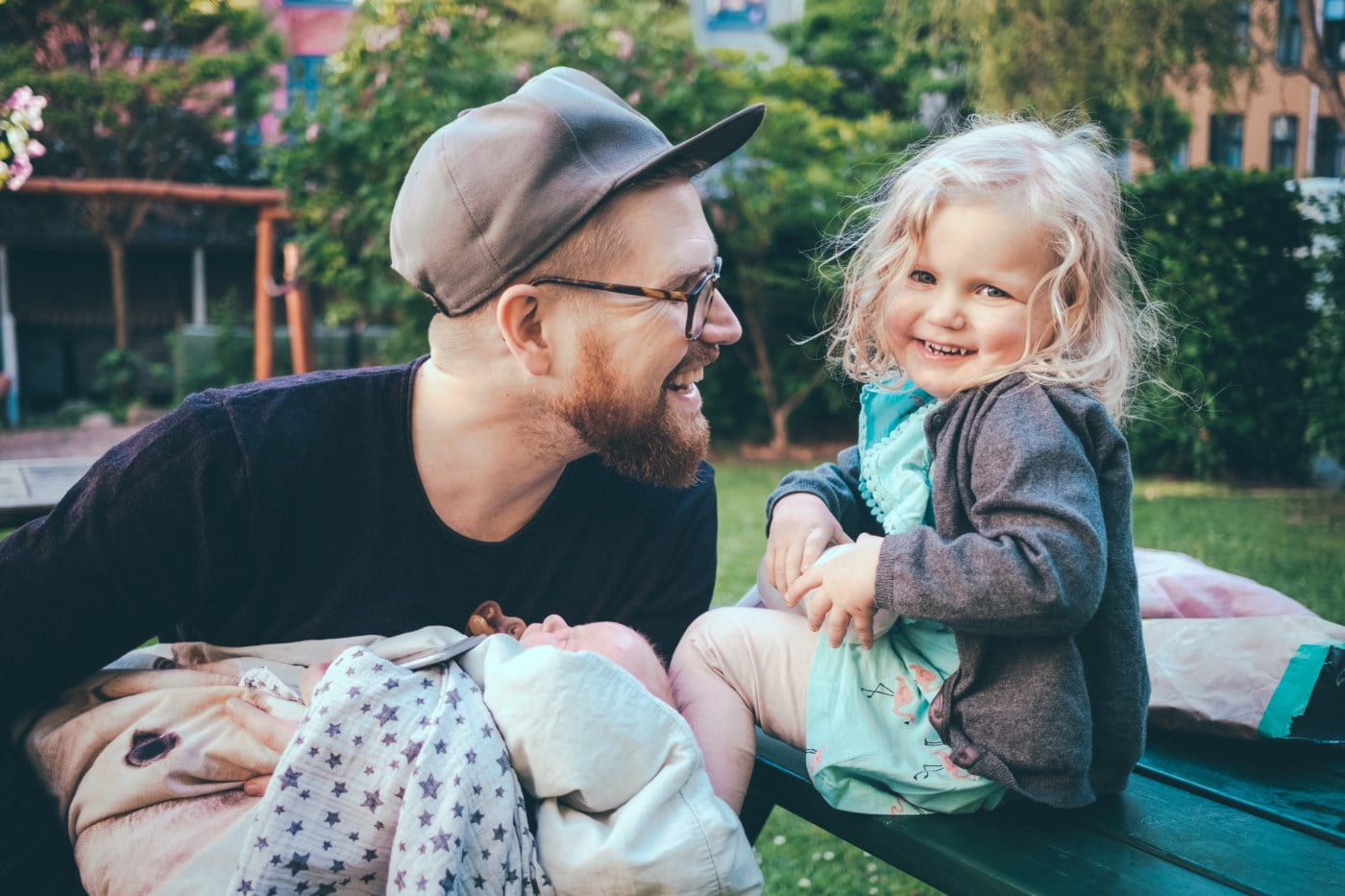 Liten jente med et stort smil som sitter ved siden av faren sin, som holder en sovende baby, i en fargerik utendørssetting.